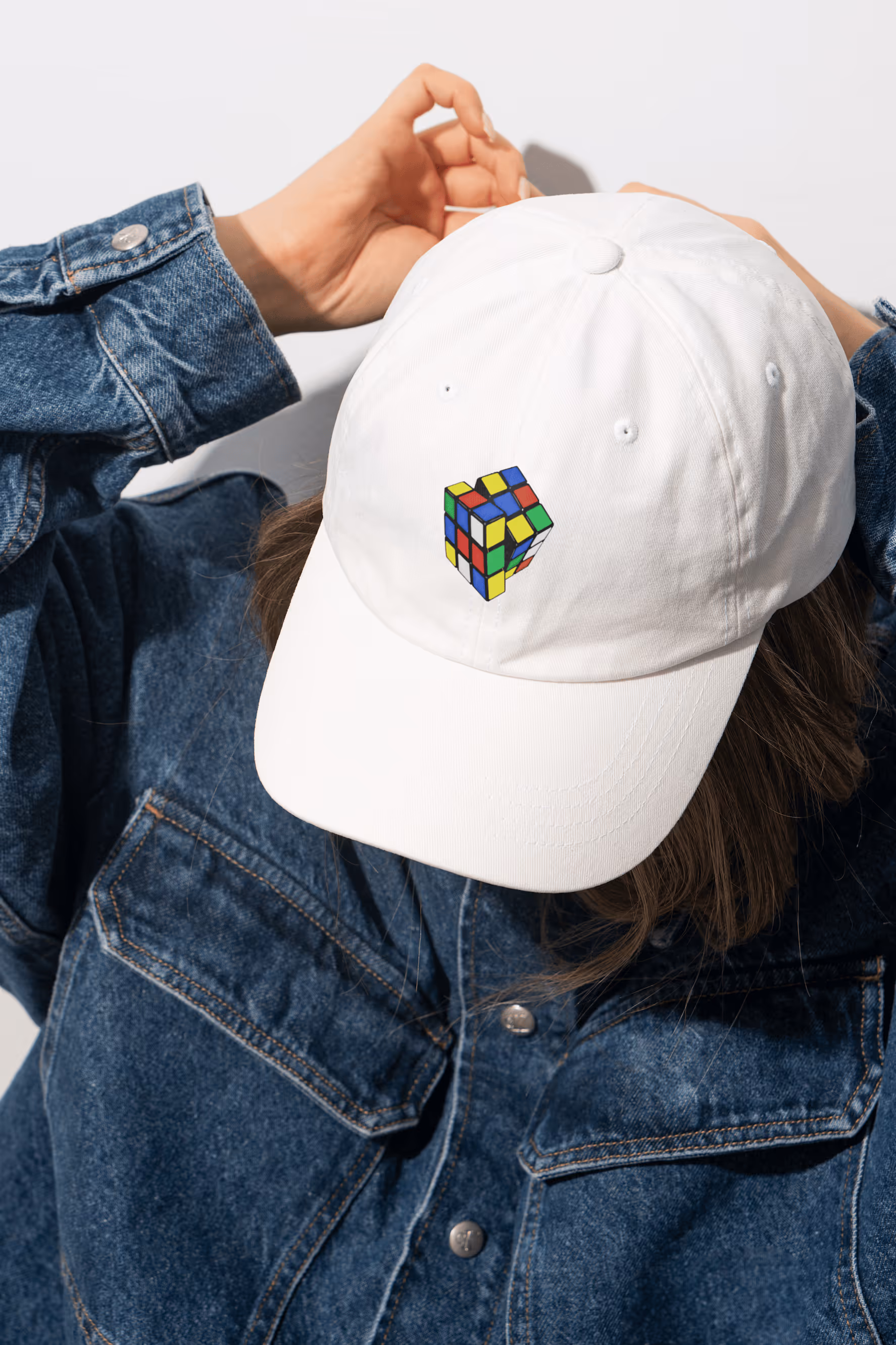Weisse Baseballkappe mit Rubik's-Cube-Stickerei, getragen von Person in Jeansjacke.