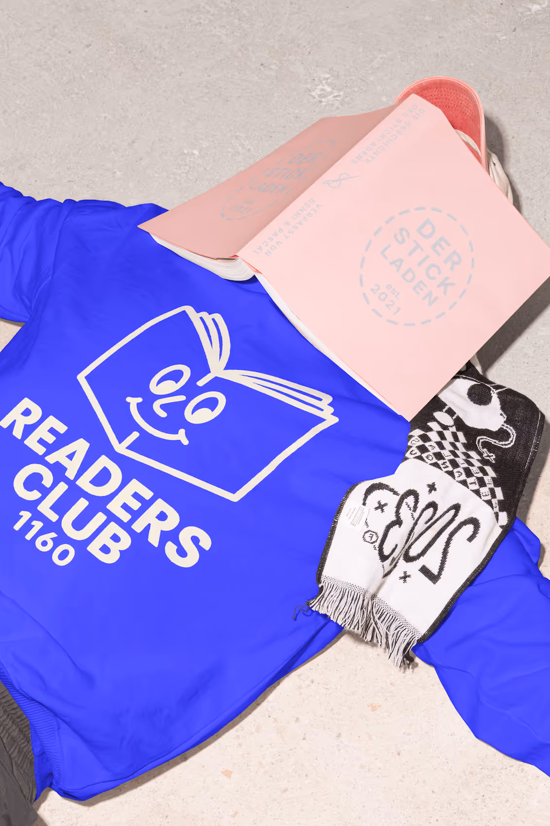 Person mit blauem Pullover mit „READERS CLUB 1160“-Aufdruck liegt am Boden mit Buch auf dem Gesicht