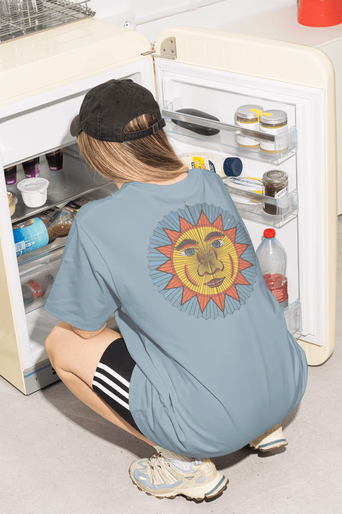 Person mit schwarzer Kappe trägt hellblaues T-Shirt mit buntem Sonnenmotiv auf dem Rücken und schaut in offenen Kühlschrank.