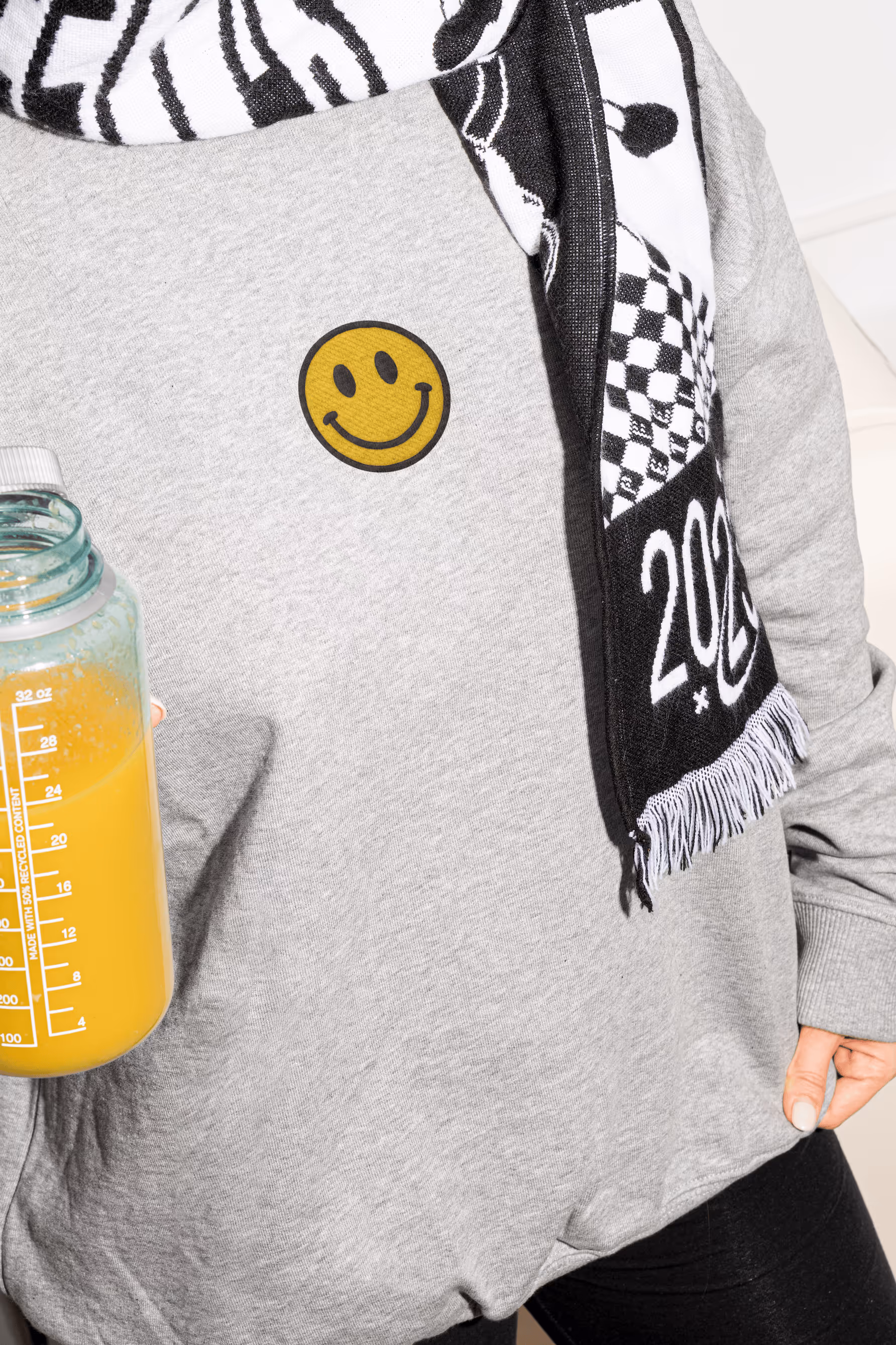 Person trägt grauen Pullover mit gelbem Smiley-Aufnäher hält eine Flasche mit Orangensaft.