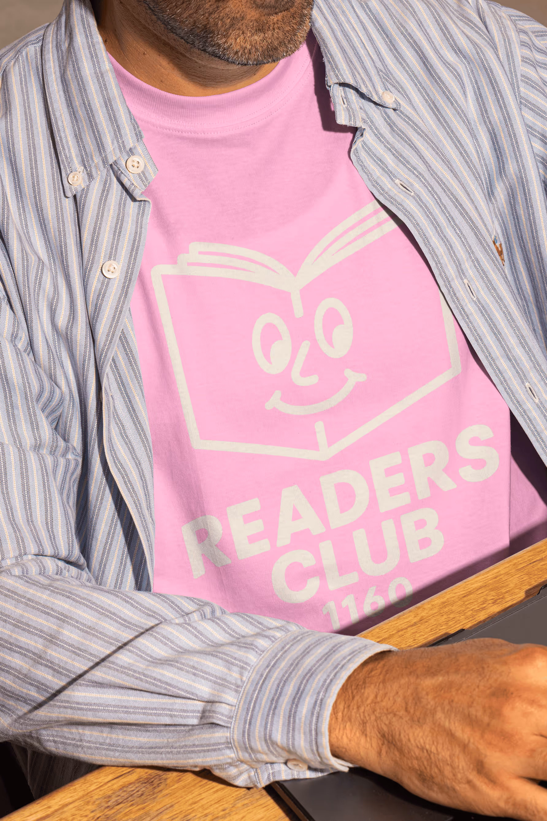 Mann trägt rosa T-Shirt mit „READERS CLUB 1160“-Aufdruck und gestreiftem Hemd beim Arbeiten.