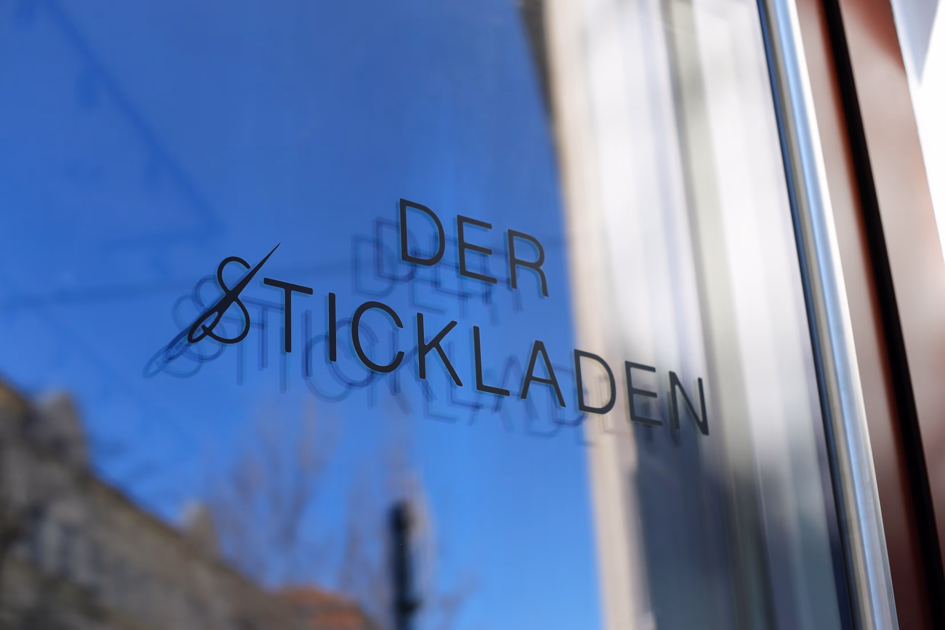Schriftzug „DER STICKLADEN“ mit Nadel-Symbol auf Glastüre, blauer Himmel spiegelt sich im Hintergrund.