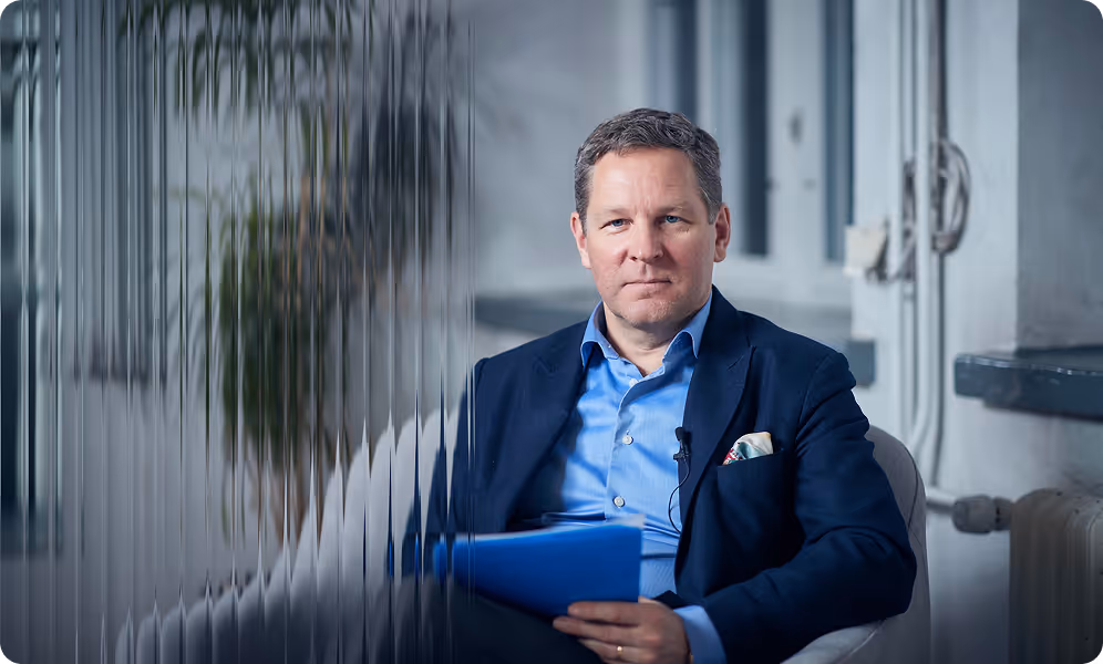 Man in a blue shirt and dark blazer sitting in a chair with folded hands holding a blue folder, separated by a textured glass panel.
