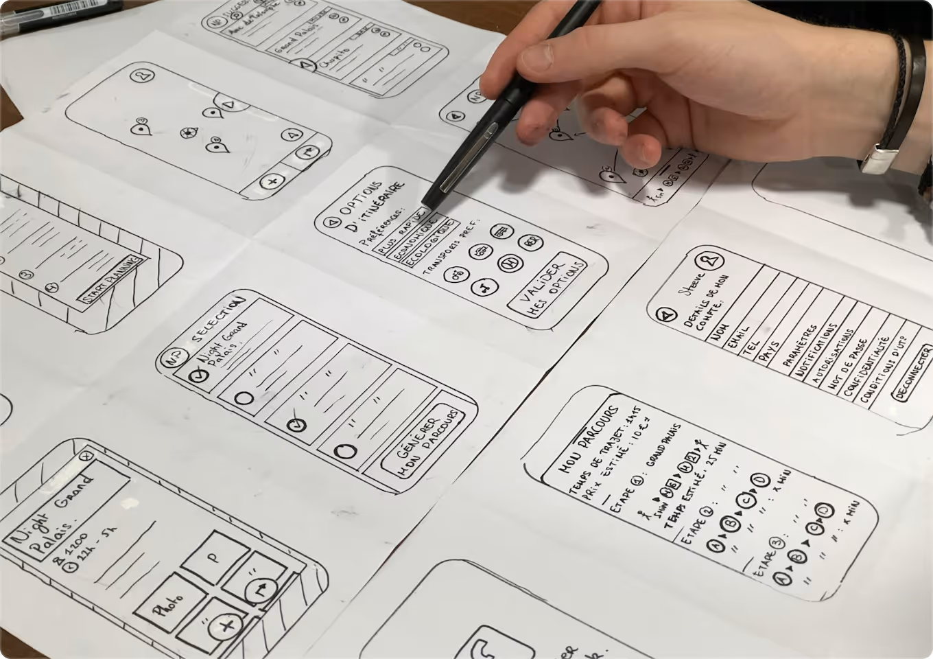A hand holding a pen pointing at sketched wireframe designs of a mobile app on paper, showing screens with maps, route options, and input forms.