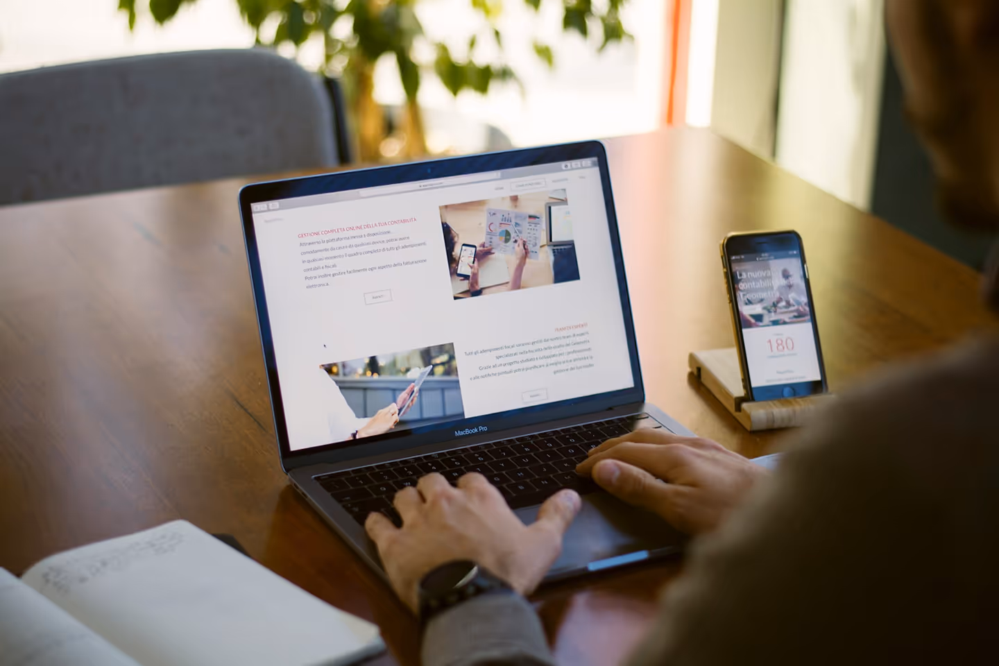 Person using a MacBook Pro on a wooden table with a smartphone on a stand beside the laptop and an open notebook nearby.