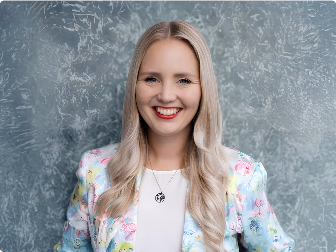 Smiling woman with long blonde hair wearing a floral blazer and a necklace with a pendant.