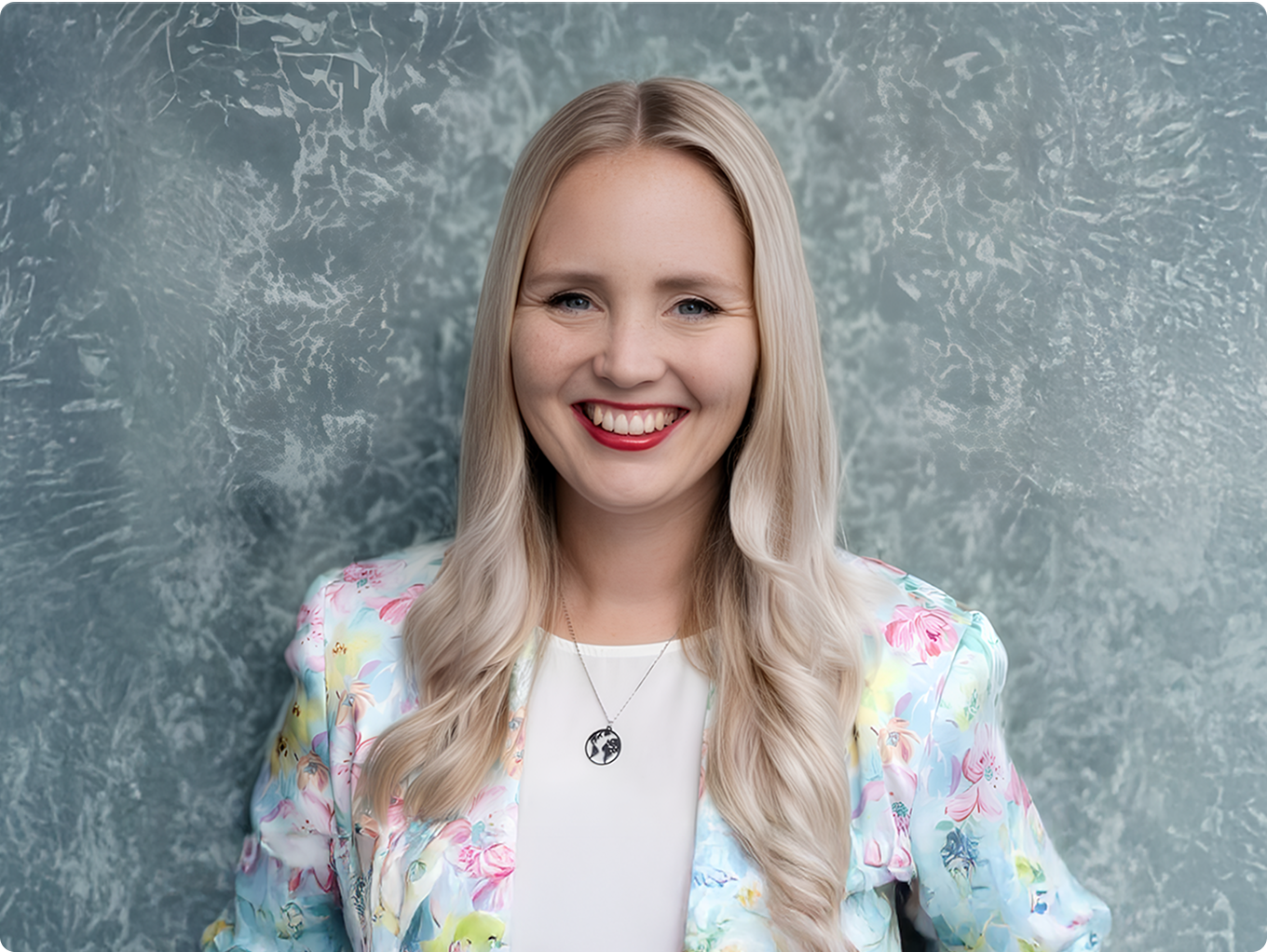 Smiling woman with long blonde hair wearing a floral blazer and red lipstick against a textured gray background.