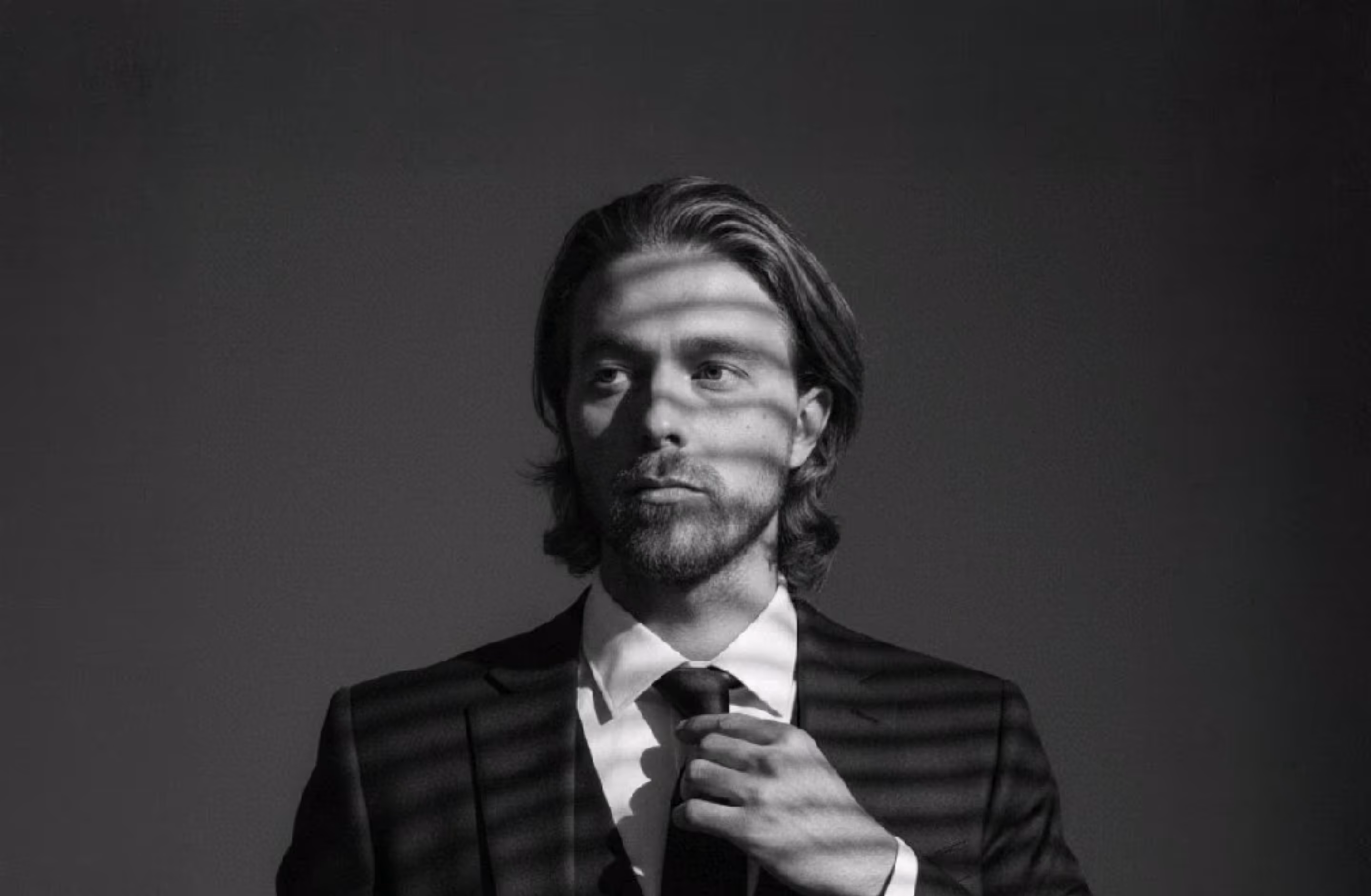 Black and white portrait of a man in a suit adjusting his tie with shadow stripes across his face.