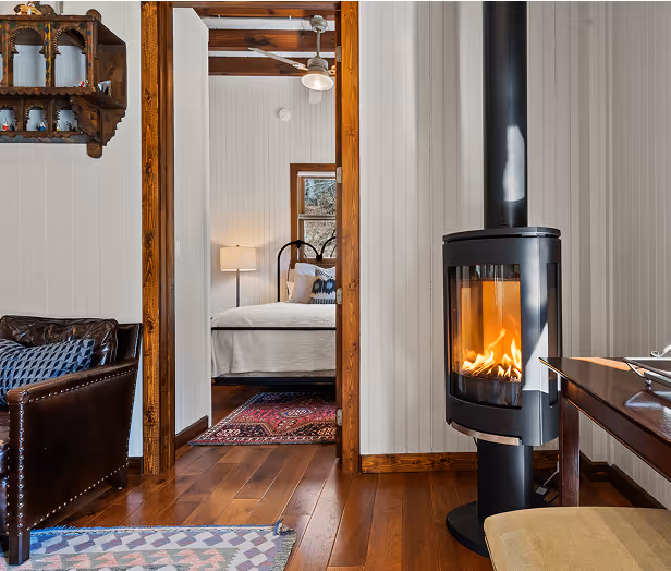 Cozy interior with a lit modern wood stove, wooden floors, leather chair, and a view into a bedroom with a bed and lamp.
