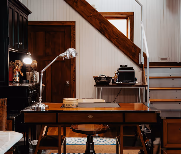Rustic home office with a wooden desk, adjustable desk lamp, chair, and staircase in the background.