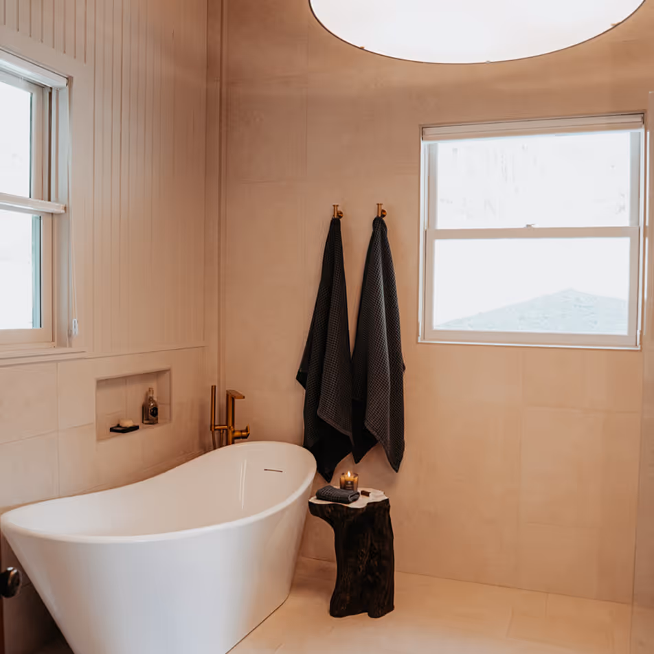 Minimalist bathroom with a white freestanding bathtub, two dark towels hanging on brass hooks, a small wooden stool with a rolled towel and lit candle, and two windows.