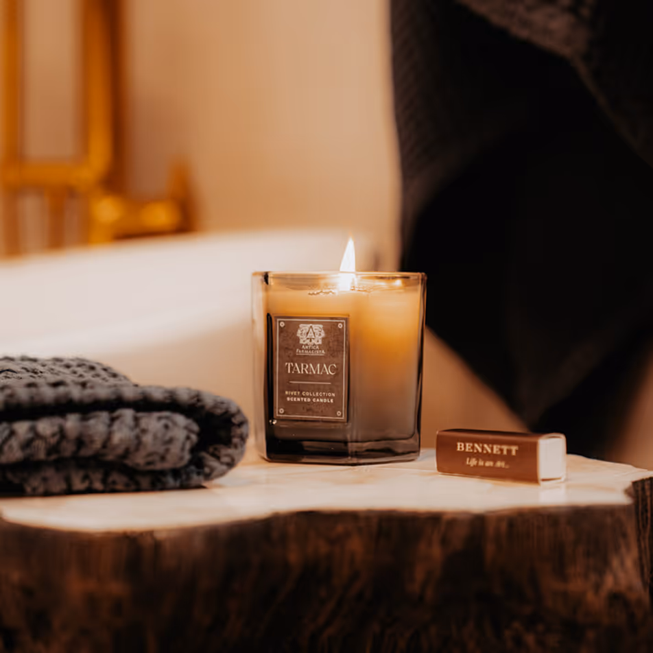 Lit Tarmac scented candle in a frosted glass container on a wooden surface next to folded dark textured fabric and a brown Bennett matchbox.