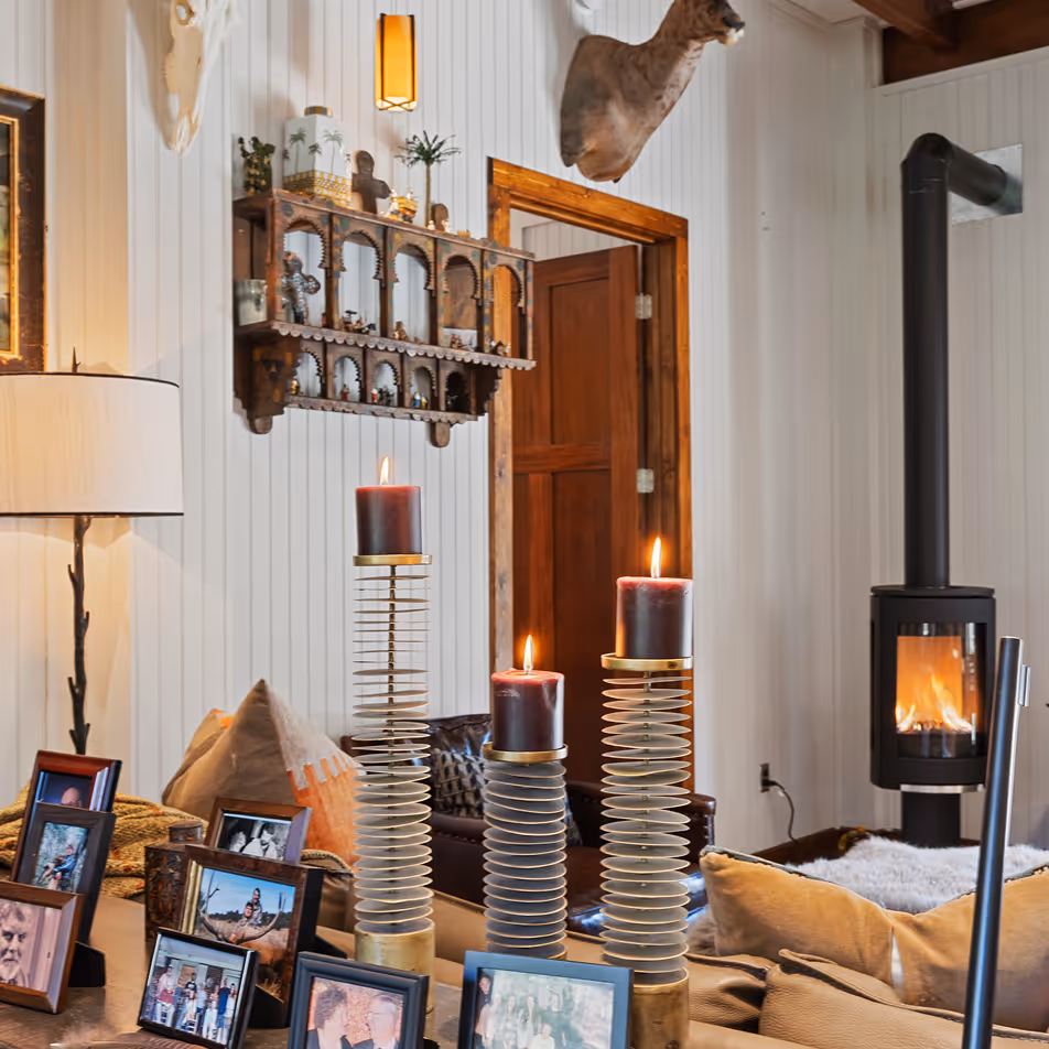 Cozy living room with lit candles on spiral metal holders, framed photos on a table, a wood-burning stove, and mounted deer head on the wall.