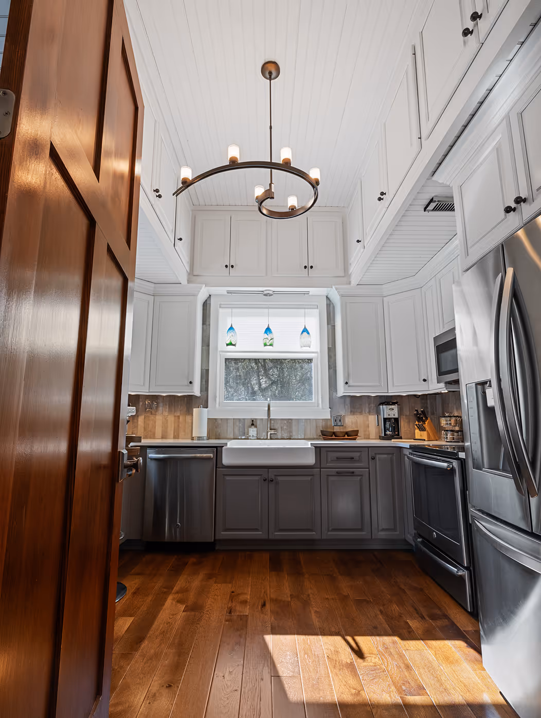 Modern kitchen with wooden floor, white upper and gray lower cabinets, a farmhouse sink, stainless steel appliances, and a unique spiral chandelier under a white paneled ceiling.