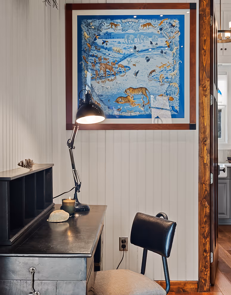 Cozy workspace with dark wooden desk, black adjustable lamp, beige cushioned chair, and framed blue wildlife illustration on white paneled wall.