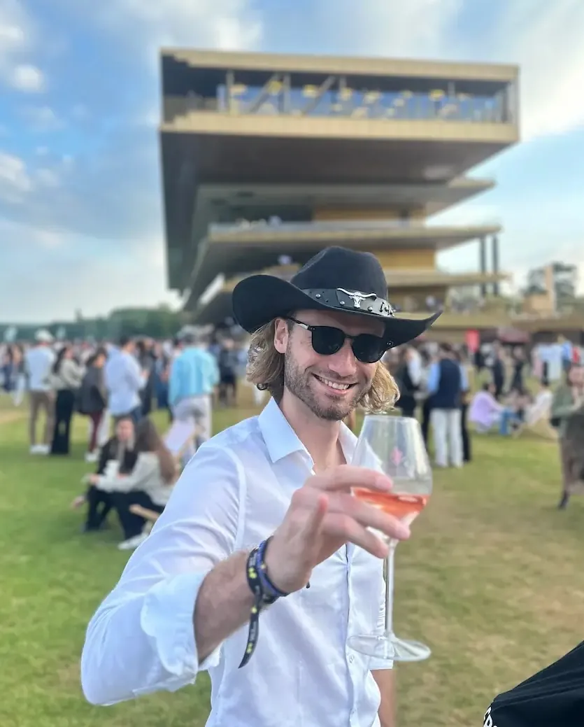 Victor BEAU brandit un verre de rosé à l'hippodrome de Longchamp. Heureux, en chemise blanche et avec son chapeau de cow-bow !