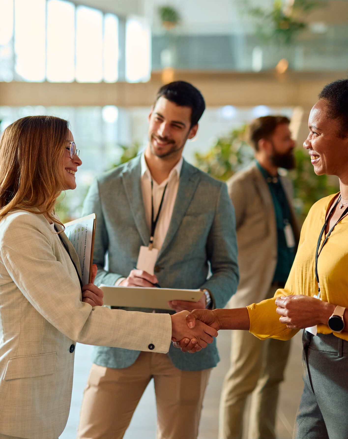 Two professional women smiling and shaking hands while two men in business attire talk in the background in a modern office setting.