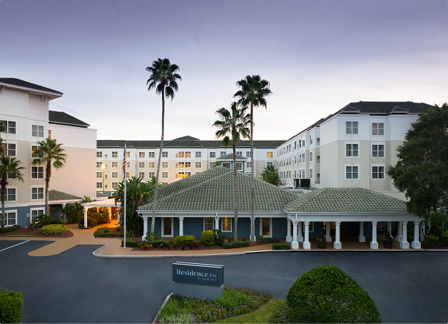 Exterior view of a Residence Inn by Marriott hotel with white and beige facade, palm trees, and a driveway entrance at dusk.