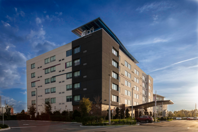 Exterior view of a modern six-story Aloft Hotels building under a partly cloudy blue sky during sunset.