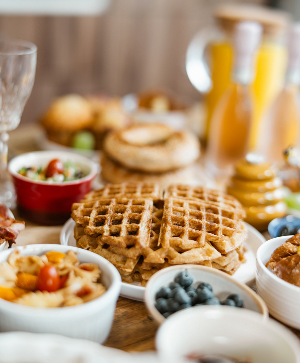 Plate of golden brown waffles surrounded by bowls of blueberries, fruit salad, and other breakfast items on a wooden table.