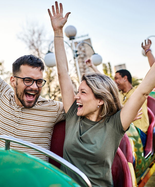 A joyful couple with arms raised, laughing while riding an amusement park roller coaster.