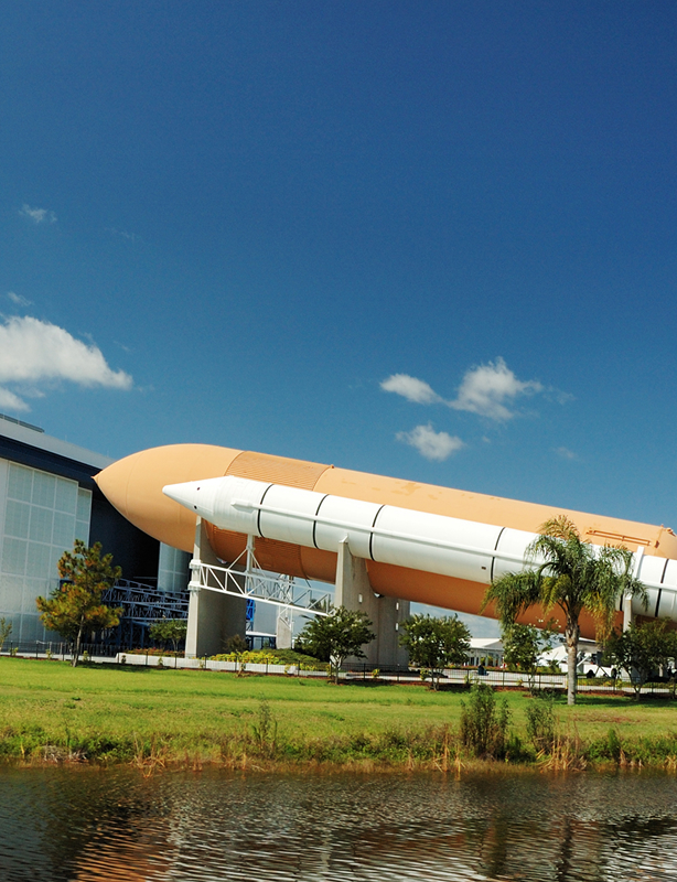 Large space shuttle external fuel tank and solid rocket boosters displayed outdoors near a building under a clear blue sky.