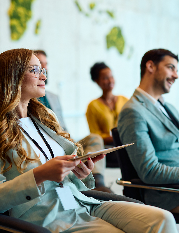 Woman with glasses holding a tablet, attentively listening in a professional meeting with colleagues in the background.