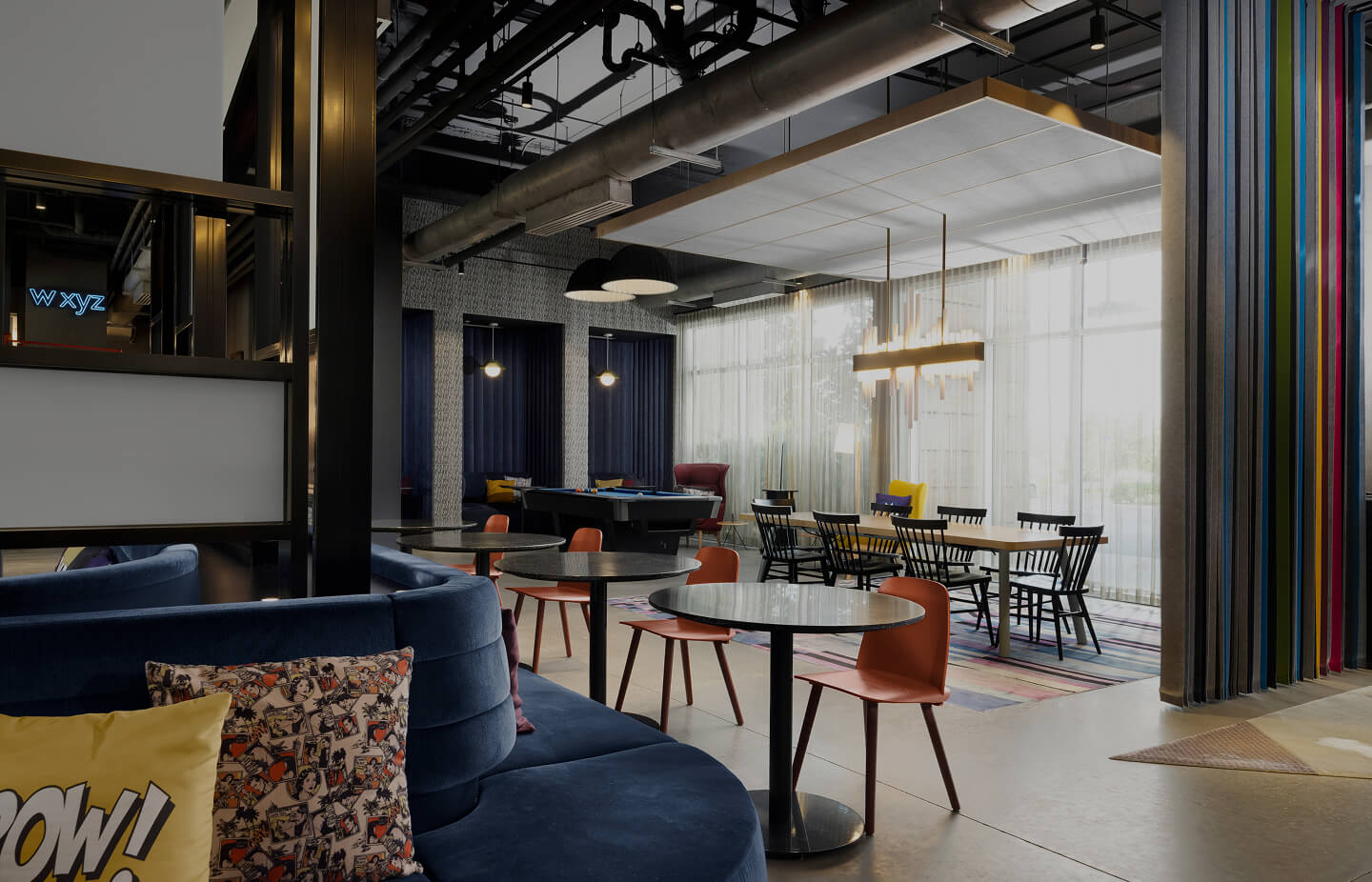 Modern lounge area with blue curved sofa, round tables with orange chairs, and a pool table near large windows with sheer curtains.