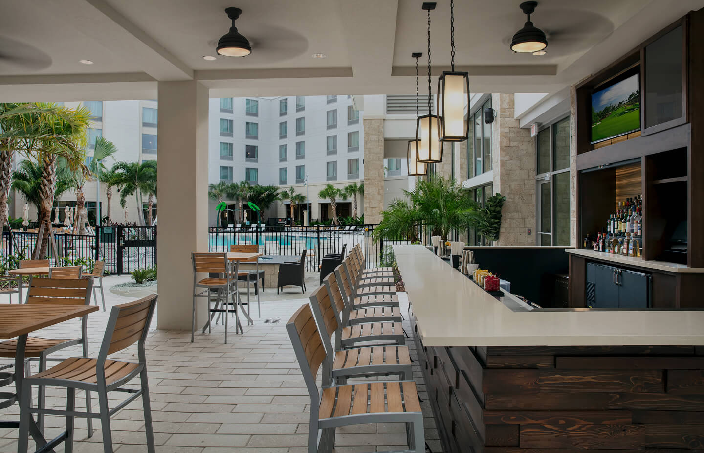 Outdoor bar area with wooden chairs, white countertop, pendant lights, and a view of a hotel pool surrounded by palm trees.