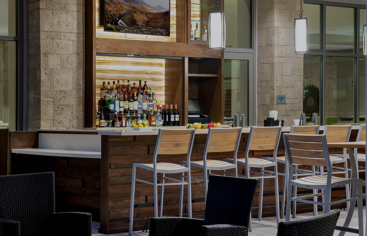 Modern indoor bar with wooden counter, bar stools, liquor bottles, and hanging pendant lights.