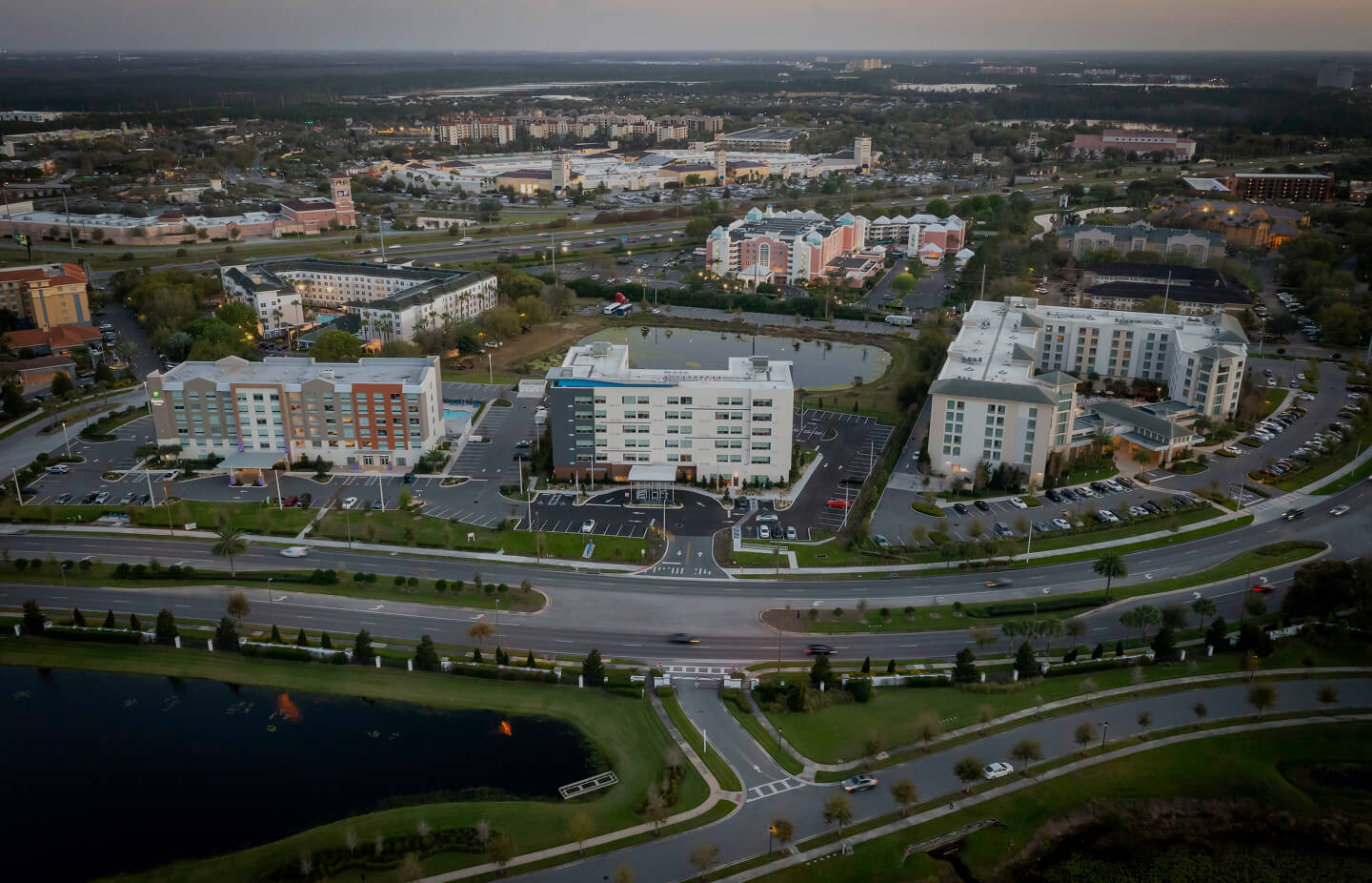 Aerial view of a suburban area at dusk featuring three large hotel buildings, parking lots, a pond, and roads with moderate traffic.