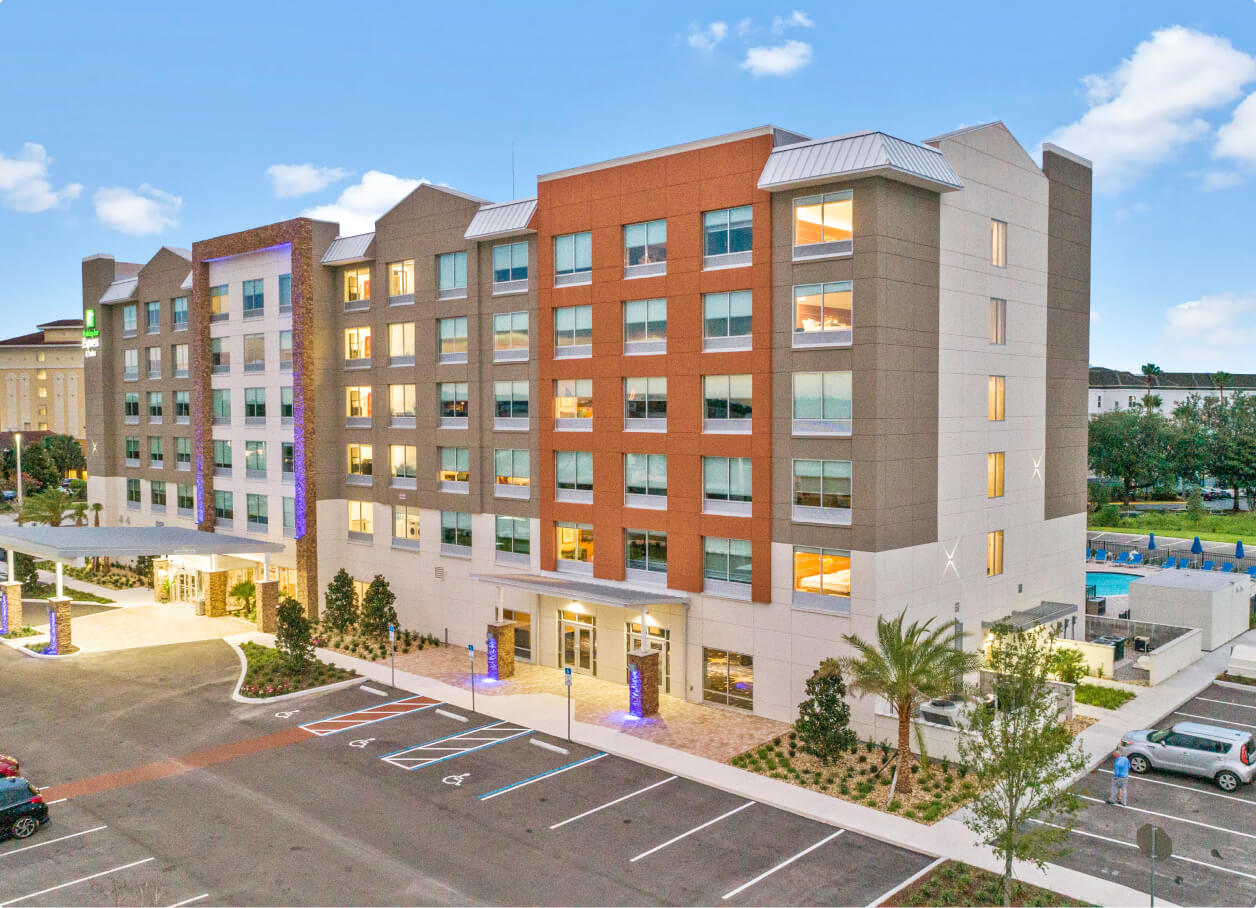 Exterior view of a modern Holiday Inn Express hotel building with a large parking lot and a swimming pool area in the background under a blue sky.