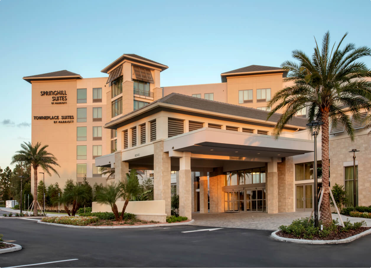 Exterior entrance of a modern hotel building with palm trees and a clear blue sky, featuring SpringHill Suites and TownePlace Suites by Marriott signage.