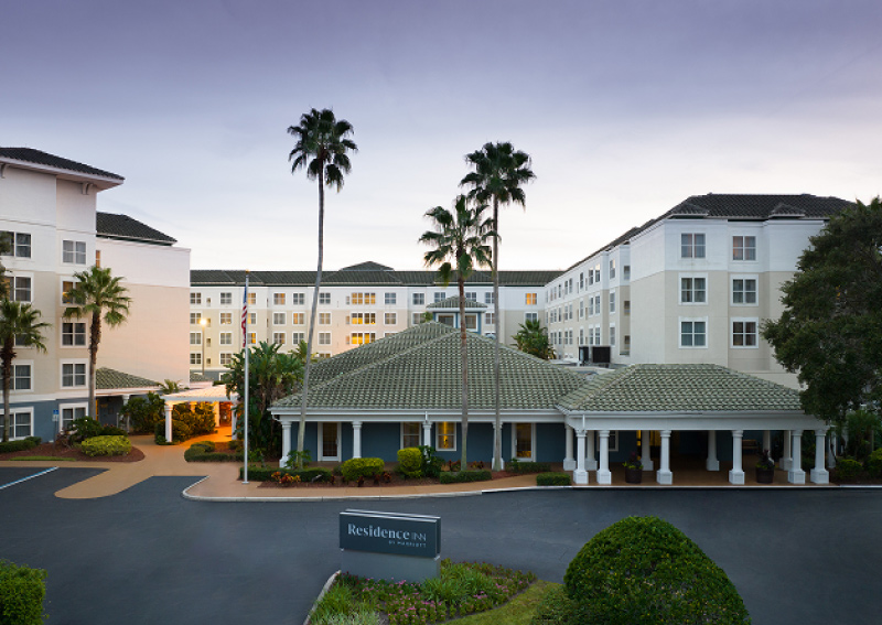 Exterior view of a Residence Inn by Marriott hotel with white and beige facade, palm trees, and a driveway entrance at dusk.