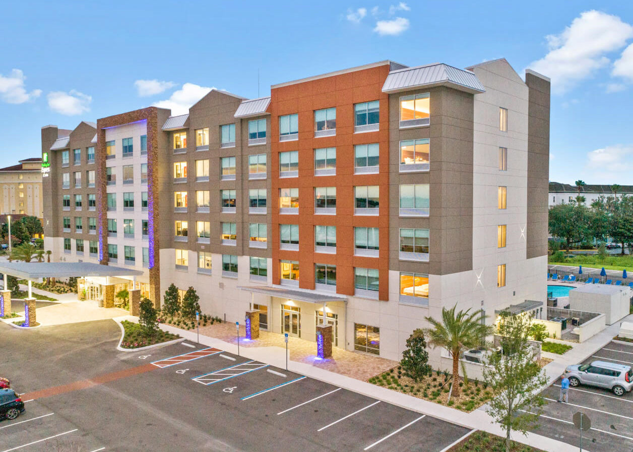 Exterior view of a modern Holiday Inn Express hotel building with a large parking lot and a swimming pool area in the background under a blue sky.
