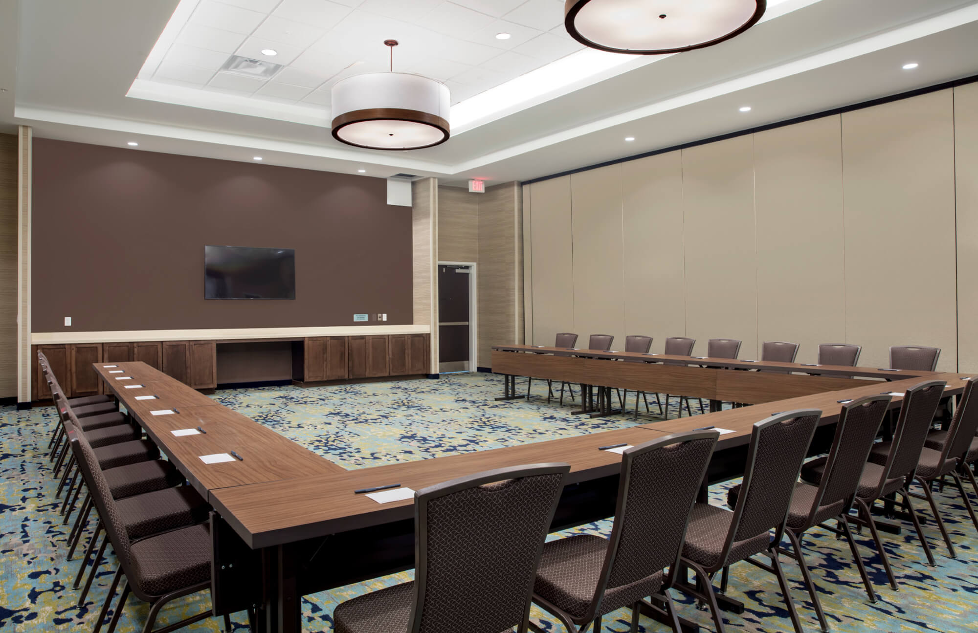U-shaped conference room with brown chairs, wood tables, carpeted floor, and wall-mounted TV screen.