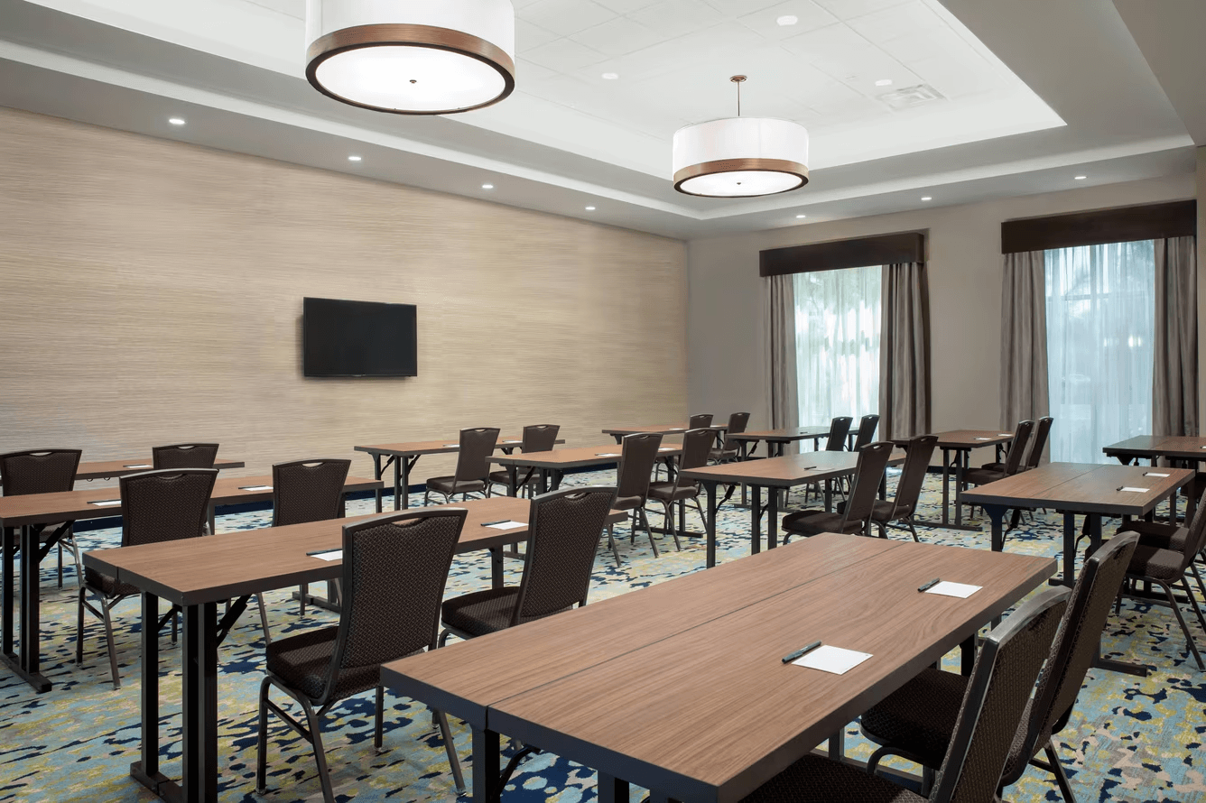 Modern conference room with rows of tables and chairs, a carpeted floor, and a wall-mounted TV.