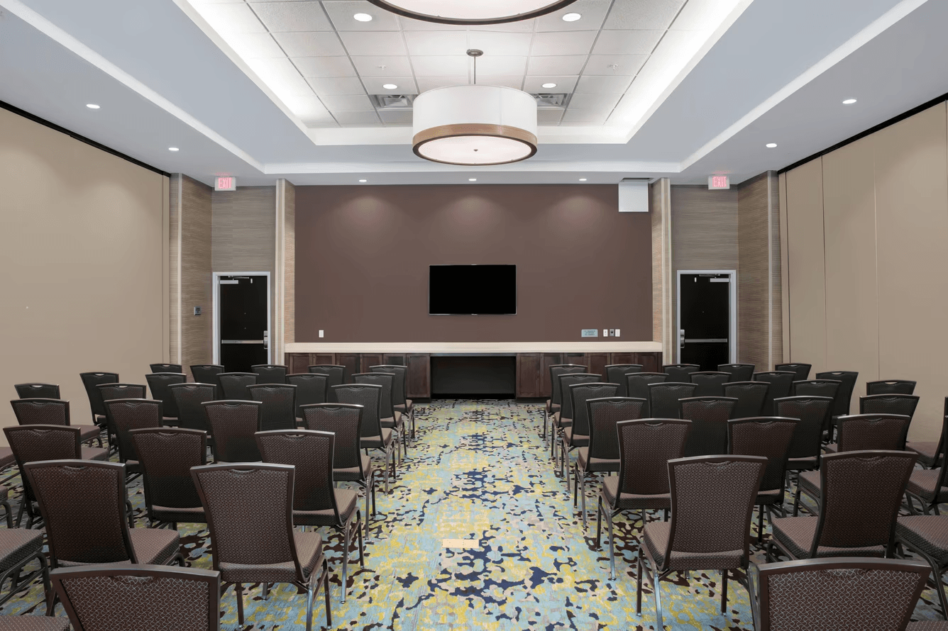 Empty conference room with rows of chairs, a patterned carpet, TV on brown wall, and ceiling light fixture.