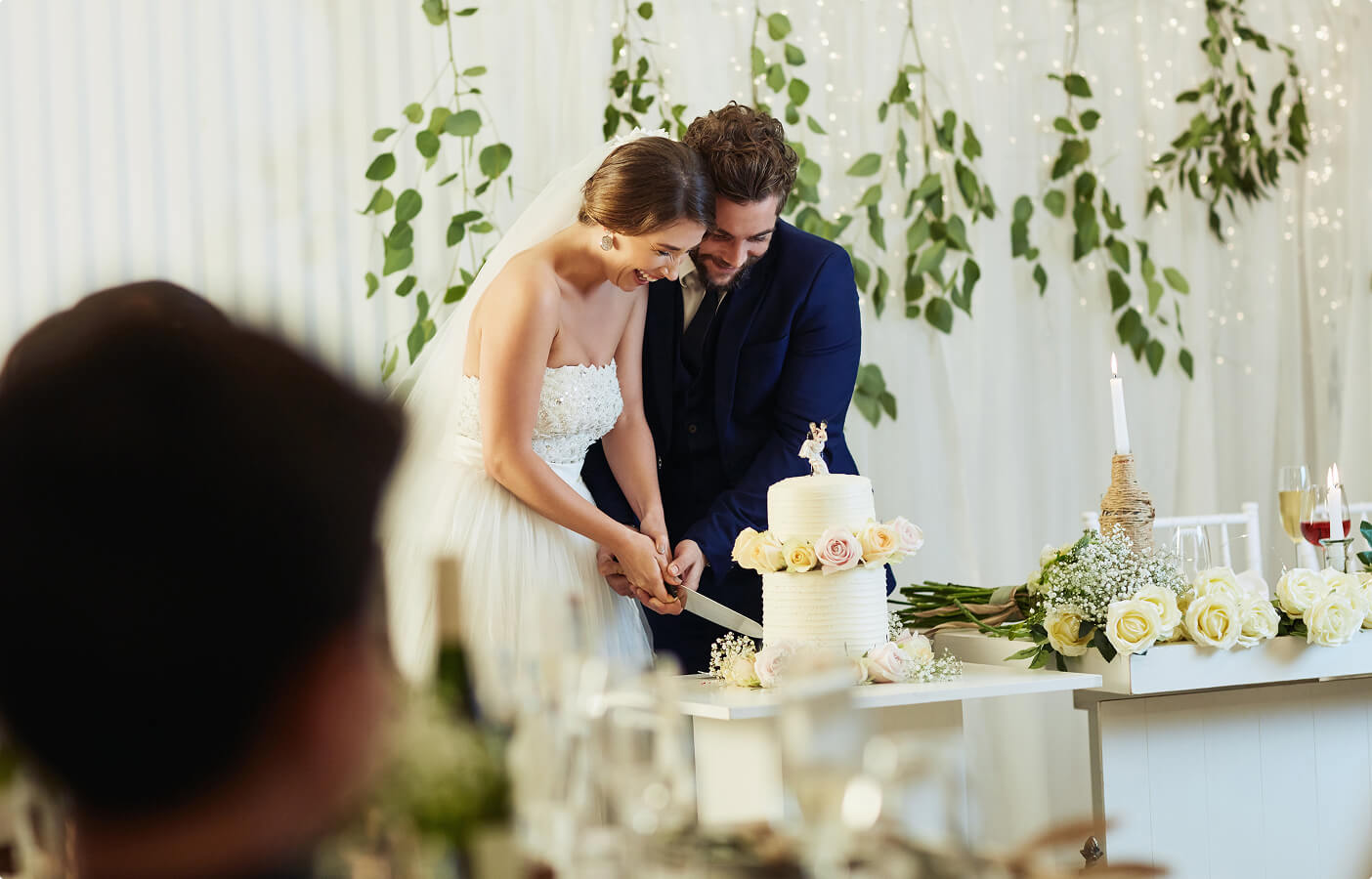 Bride and groom smiling together cutting a white, two-tiered wedding cake decorated with roses.