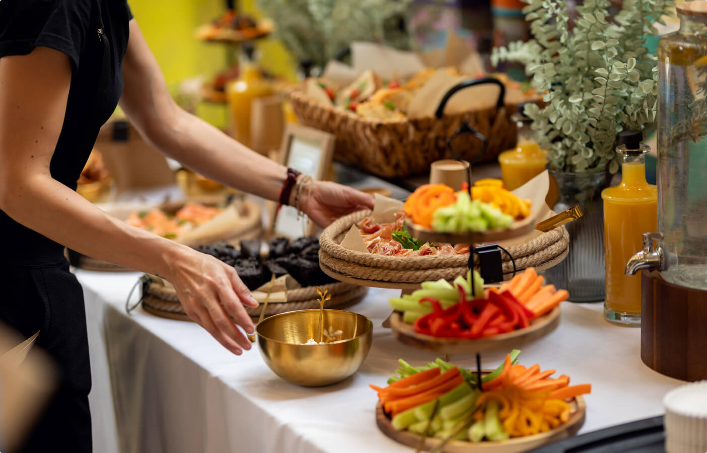 Person arranging a buffet table with plates of fresh vegetables, meats, and orange juice dispensers.