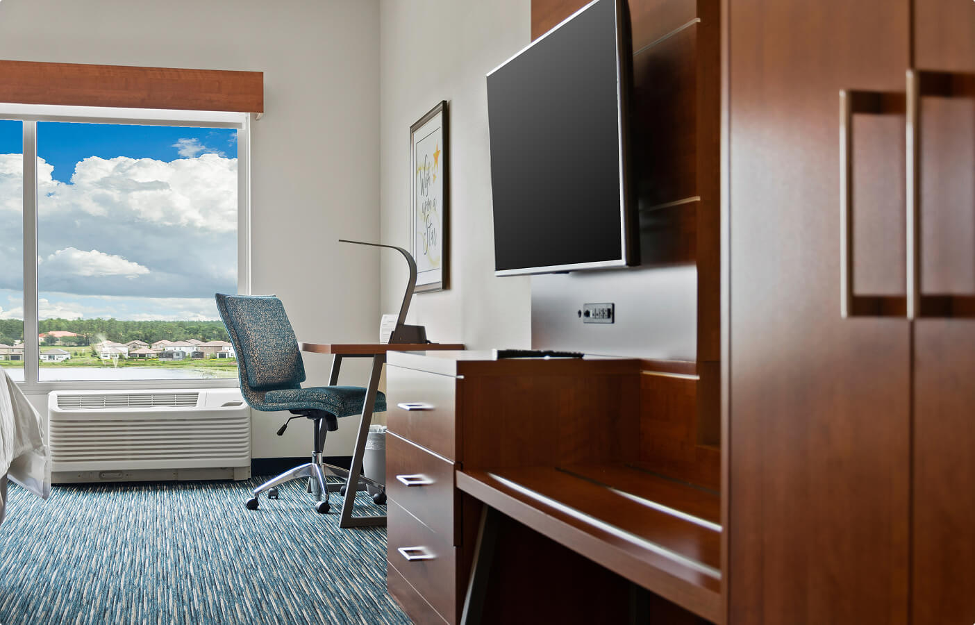 Modern hotel room workspace with blue chair, wooden desk, flat-screen TV, and large window with view.
