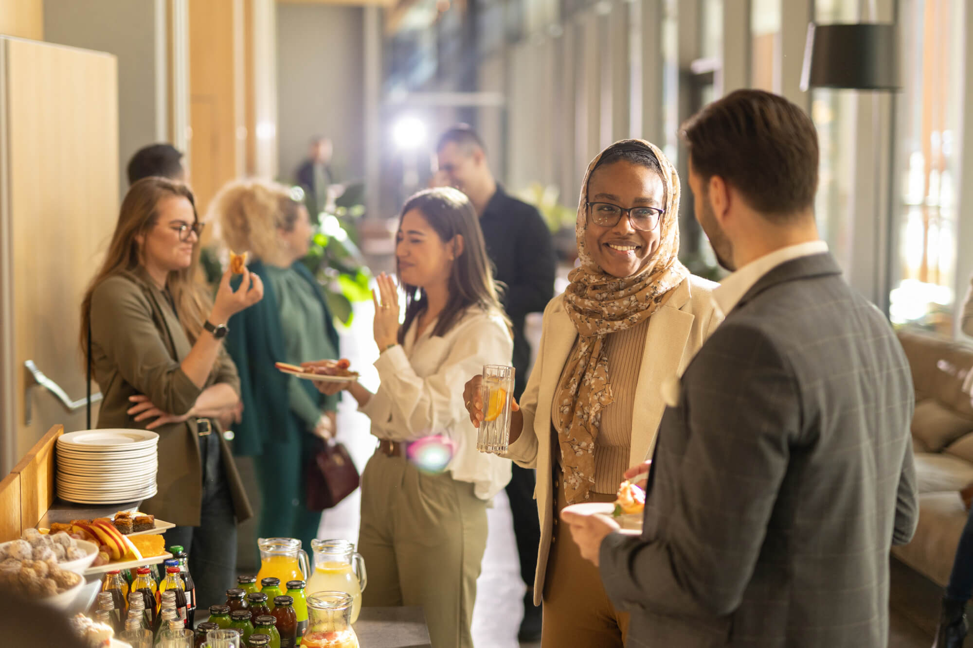 People smiling and chatting while holding drinks and plates of food at a casual indoor gathering.