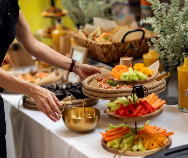 Person reaching toward a golden bowl on a table with trays of vegetables, sandwiches, and juice bottles.