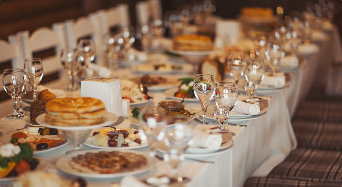 Long dining table set with white tablecloth, glassware, plates of food, and folded napkins.
