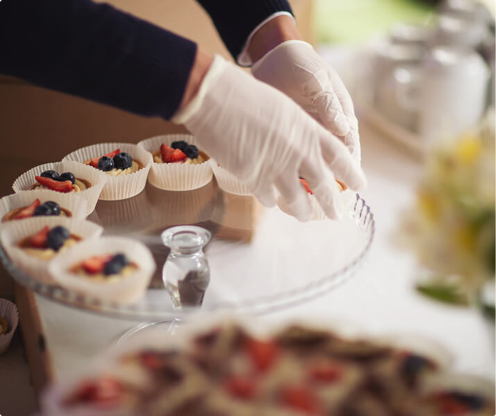 Hands in gloves arranging fruit tarts with strawberries and blueberries on a glass serving tray.