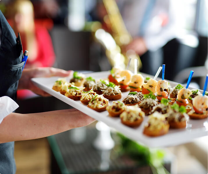 Server holding a tray of assorted savory appetizers with shrimp and garnishes on skewers.