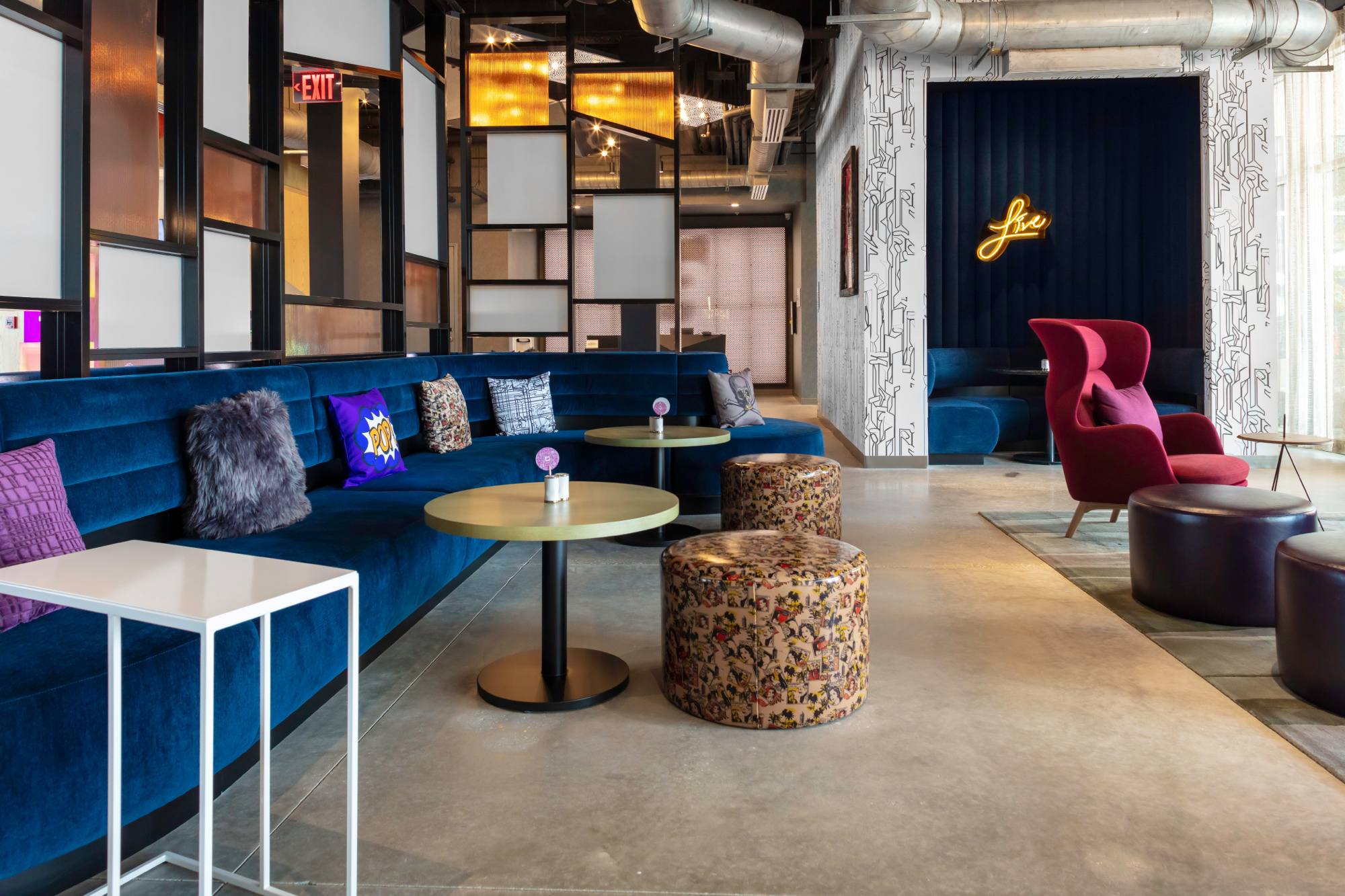 Modern lounge with blue velvet sectional, colorful cushions, round tables, patterned stools, and red armchairs.