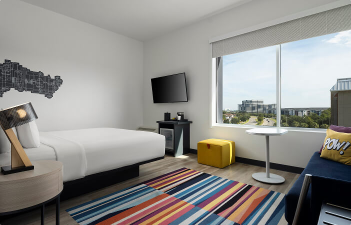 Modern hotel room with a white bed, colorful striped rug, wall TV, yellow ottoman, and large window view.