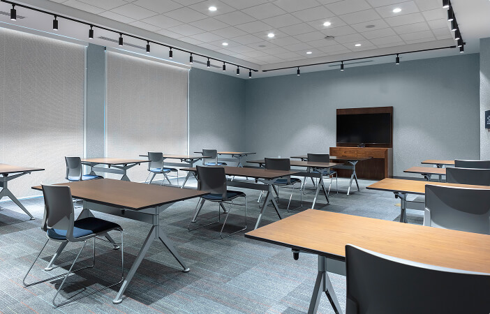 Empty modern conference room with tables, chairs, ceiling lights, and a TV on a wooden stand.