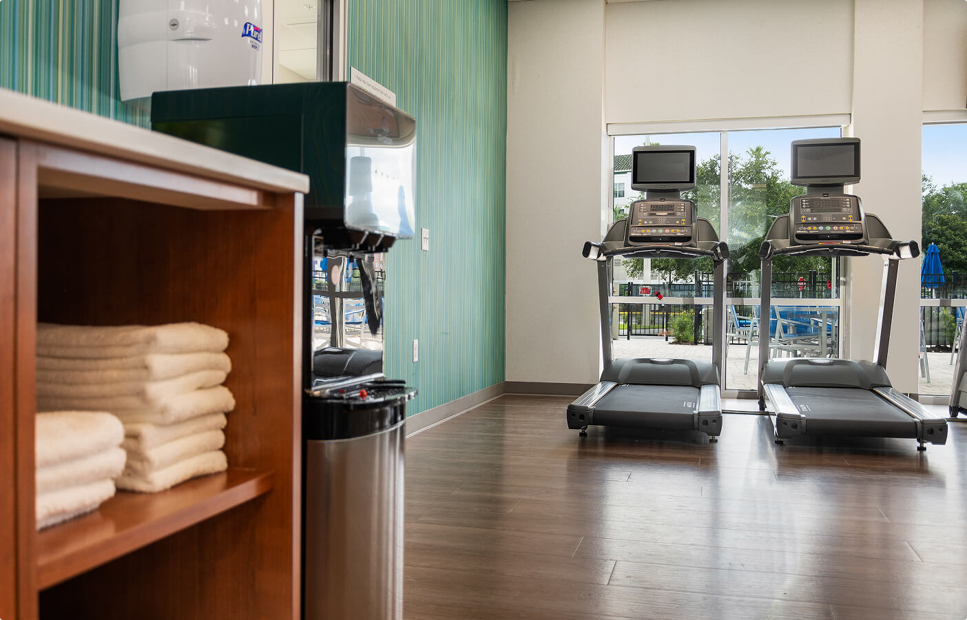 Two treadmills facing large windows with an outdoor view and a shelf with folded towels.